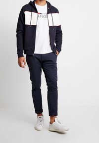 Felpa navy con strisce orizzontali bianche e rosse, abbinata a pantaloni slim-fit navy e sneakers bianche. Il tessuto ha una consistenza liscia.