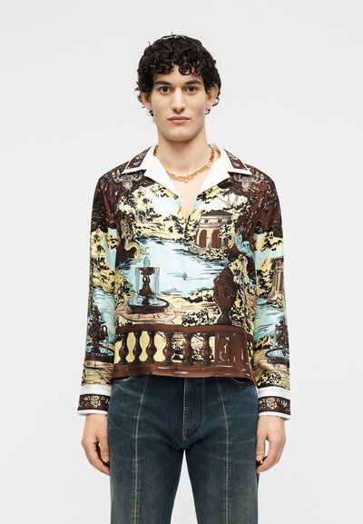 Άνδρας με σγουρά σκούρα μαλλιά, φορώντας κουμπωτό πουκάμισο με λεπτομερή τοπικό σχέδιο και σκουρόχρωμο τζιν, κοιτάζοντας μπροστά με ουδέτερη έκφραση.