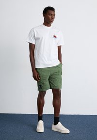 Balti marškinėliai su raudonų vyšnių grafika, žali krovinių šortai ir kreminės spalvos sportbačiai. Šortai turi šonines kišenes ir juodą etiketės lopelį.