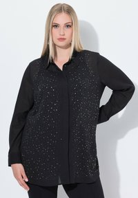 Schwarzes Hemd mit langen Ärmeln, aus leicht durchscheinendem Stoff, verziert mit einem Muster aus silbernen Verzierungen auf der Vorderseite.