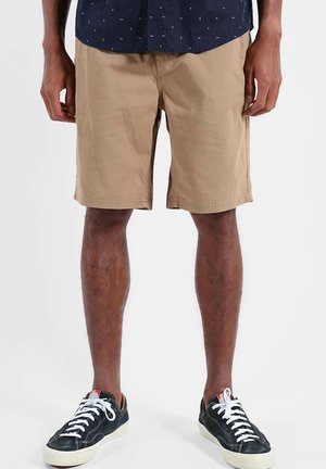 Homme portant un short beige jusqu'aux genoux, des baskets noires basses avec des lacets blancs, et une partie d'une chemise bleu foncé à motif.