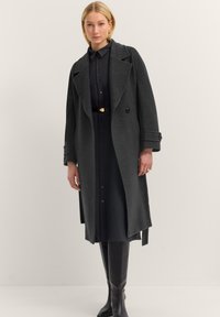 Frau in einem dunkelgrauen langen Mantel über einem schwarzen knöpfbaren Kleid, schwarzem Gürtel, goldenen Ohrringen und kniehohen schwarzen Stiefeln vor einem schlichten Hintergrund.