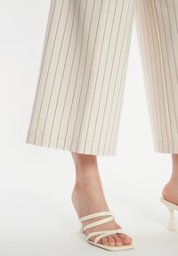 Beige weite Hosen mit schwarzen vertikalen Streifen, die mit einem Saum enden, der knapp über dem Knöchel liegt. Kombiniert mit weißen offenen Sandalen mit Absatz.