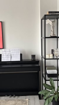 Czarny cyfrowy fortepian z nutami, czarny świecznik oraz czarna metalowa półka z różnymi przedmiotami, w tym rośliną i statuą.