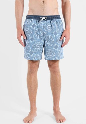Homme portant un short de bain bleu avec des motifs géométriques circulaires blancs et une ceinture élastique bleu marine, debout pieds nus sur fond blanc.