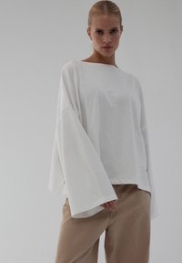 Biały bawełniany top z szerokimi, rozkloszowanymi rękawami i dekoltem typu łódka. Do tego beżowe spodnie, gładka faktura, minimalistyczny styl.