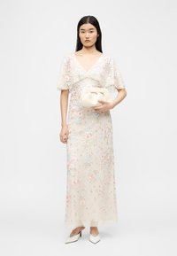 Γυναίκα με διαφανές λευκό μακρύ φόρεμα με floral μοτίβο και φουσκωτά μανίκια, κρατώντας ένα μαλακό λευκό τσαντάκι, φορώντας λευκές μυτερές γόβες.