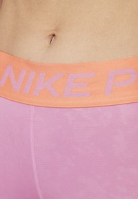 Ružové atletické šortky so širokým oranžovým pásom s nápisom "NIKE" v svetlo fialovej farbe. Hladká textúra, priliehavý dizajn a silné šitie.