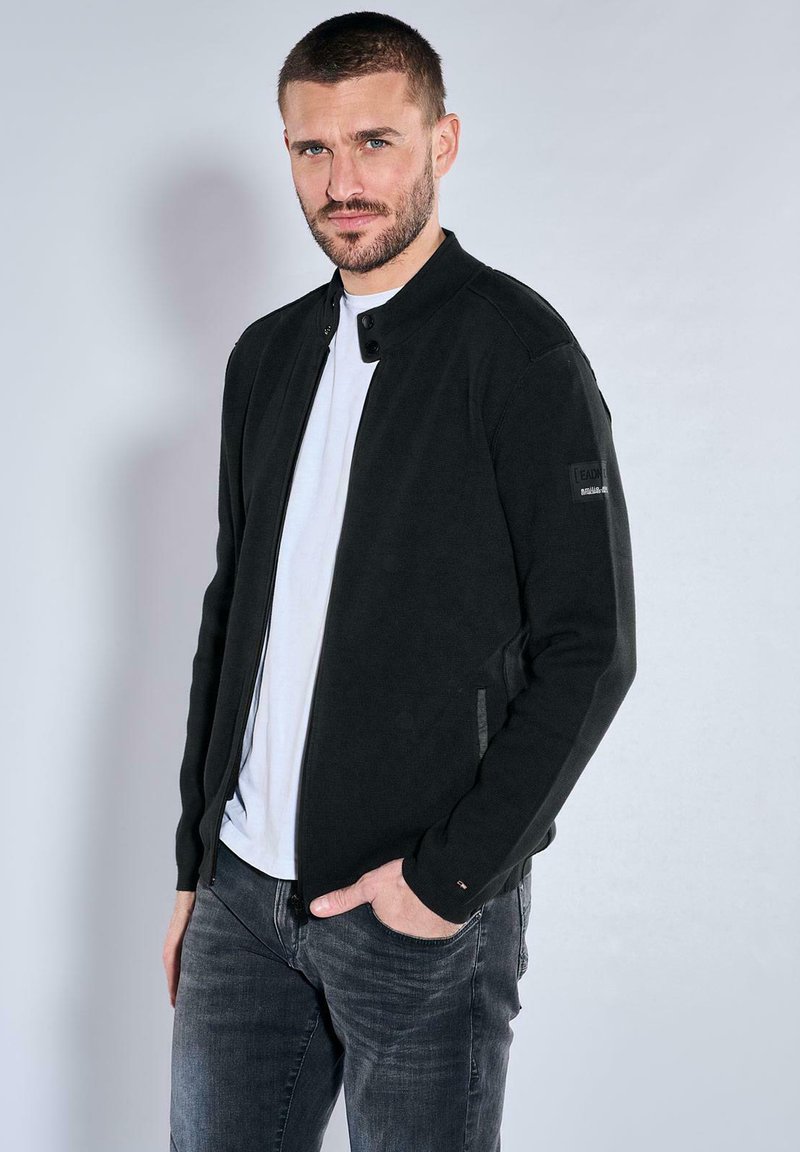 Black bomber jacket made of soft fabric with a zip front, snap closure, side pockets, and subtle logo details on sleeves. Paired with gray jeans.