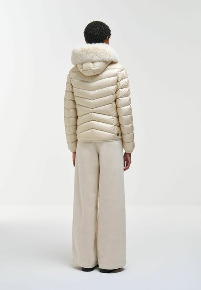 Personne aux cheveux courts et bouclés, portant une doudoune crème avec capuche en fourrure et un pantalon beige à jambes larges, debout de dos sur fond blanc.