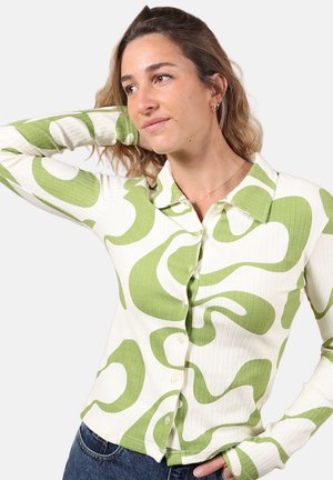 Femme aux cheveux ondulés portant une chemise boutonnée à manches longues avec des motifs abstraits verts et un jean bleu, posant avec une main derrière la tête.
