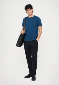 Blå t-shirt med korta ärmar, svarta byxor och svart läderväska. Mannen står avslappnat och bär svarta skor. Enkel design.