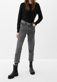 Personne portant un pull noir texturé, un jean gris taille haute avec revers, des bottines noires épaisses, debout devant un fond blanc uni.