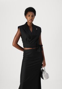 Svart stripete cropped blazer med knytet, kombinert med et svart blyantskjørt. Modellen holder en sølvfarget satinhåndveske.