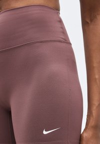 Κοντινό πλάνο ατόμου που φορεί αθλητικά σορτς Nike σε βυσσινί υψηλής μέσης, δείχνοντας την υφή του υφάσματος και το λογότυπο στον μηρό.