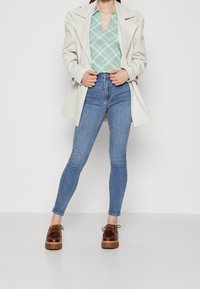 Beżowy trench coat nałożony na koszulę w zielono-niebieską kratę, w połączeniu z niebieskimi dżinsami o wysokim stanie i brązowymi butami na platformie z detalem sznurowania.