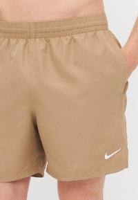 Shorts de sport beiges avec une taille élastique, des poches latérales et un logo Nike blanc sur la jambe gauche. Texture de tissu lisse.