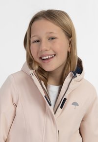 Šviesiai rožinė striukė su kapišonu, turinti tamsiai mėlyną pamušalą ir mažą pilką skėčio logotipą. Užtrauktukas su juodu apvadu ir minkšta medžiagos tekstūra.