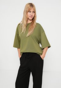 Olivengrønn oversized t-skjorte med vide korte ermer, laget av mykt stoff, kombinert med løse svarte bukser med sidelommer.