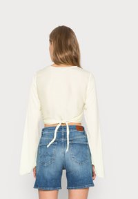 Camisola de mangas longas amarelo claro com as costas atadas; combinada com calções de ganga azuis com um remendo em pele e bolsos traseiros.