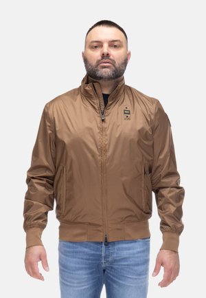 Uomo con capelli corti scuri e barba che indossa una giacca marrone con zip e jeans blu, in piedi contro uno sfondo chiaro e uniforme.