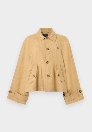 Γυναίκα που φορά καφέ μπουφάν, ανοιχτό μπλε πουκάμισο, λευκό παντελόνι, λευκά αθλητικά παπούτσια και καπέλο καφέ χρώματος, κρατώντας μια μικρή λευκή τσάντα.