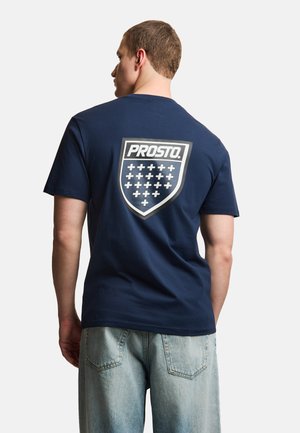 Mężczyzna noszący granatową koszulkę z logo w kształcie tarczy i napisem „PROSTO.” oraz wieloma znakami plus na plecach, połączoną z jasnoniebieskimi dżinsami.