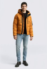 Orangene gesteppte Jacke mit schwarzem Reißverschluss und Details aus Metall, mit Kapuze und Fronttaschen, getragen über einem schwarzen Hemd und blauen Jeans.