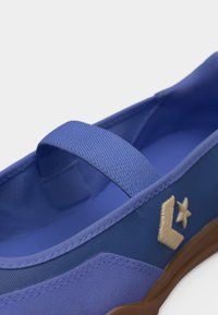 Chaussure bleu à enfiler avec une bande élastique sur le dessus, logo étoile beige et chevron sur le côté, et semelle en caoutchouc marron.