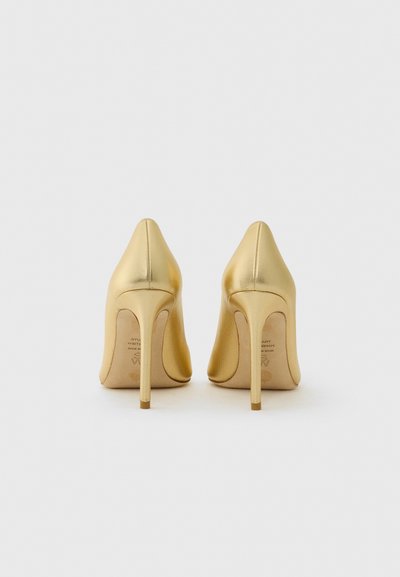 Une paire de chaussures à talons hauts en métal doré vues de l'arrière sur un fond blanc uni.