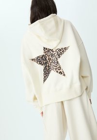 Krämfärgad munkjacka med en stor leopardmönstrad stjärna på baksidan. Mjuk tyg, oversized passform, och en känguruficka på framsidan.