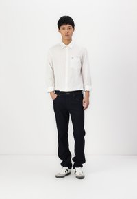 Witte, lange mouw knoopshirt met een borstzak, gecombineerd met donker denim jeans en witte sportschoenen met grijze accenten.