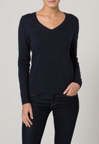 Marineblauer V-Ausschnitt-Pullover aus weichem Strickmaterial. Lange Ärmel, taillierte Silhouette und gerippter Saum. Kombiniert mit dunkelblauen Denim-Jeans.