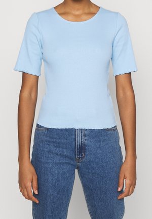 Personne portant un t-shirt bleu clair à manches courtes côtelé avec des bords festonnés et un jean en denim bleu sur un fond uni.