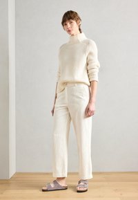 Crème ribgebreide turtleneck trui, gecombineerd met crème wijde broek en lila instapsandalen met zilveren accenten. Houten vloer als achtergrond.