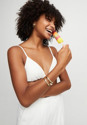 Top de bikini blanco, pulseras doradas, sosteniendo una paleta de colores. Pelo rizado, sonriendo, con piel suave y pantalones de tela ligera.