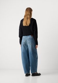 Suéter negro texturizado combinado con jeans de mezclilla azul sueltos que presentan grandes bolsillos y un parche marrón contrastante en la parte posterior. Zapatos negros.