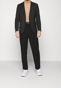 Homme portant une veste de costume noire et un pantalon noir, un pull col roulé beige, une ceinture noire et des sneakers blanches, se tenant contre un fond blanc.