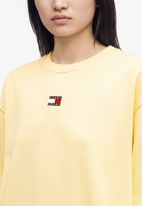 Jeune personne aux cheveux raides et foncés portant un sweat-shirt jaune pâle avec un petit logo Tommy Jeans sur la poitrine