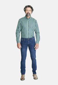 Camisa de cuadros verde y azul oscuro, jeans azules y zapatos beige. El modelo está de pie con los brazos relajados a los lados.