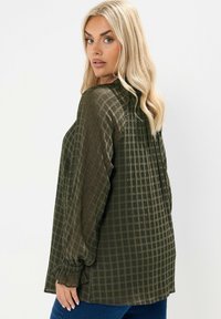 Bluzka w zielono-oliwkowe kraty, lekki materiał, długie rękawy z elastycznymi mankietami, luźny krój, zaokrąglony dół, subtelna tekstura.