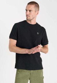 T-shirt noir à manches courtes en tissu texturé avec un col rond et un petit logo circulaire sur la poitrine gauche.