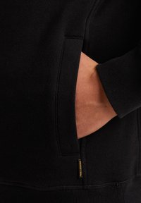 Black sweatshirt featuring a side pocket and textured fabric. Includes a small yellow brand tag at the hem, highlighting its design.