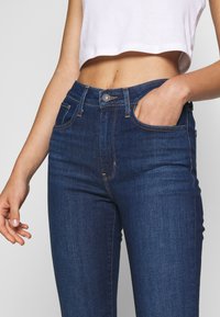 Jeans de mezclilla oscura, de talle alto y ajustados, con bolsillos frontales y cierre de botón de metal. Textura suave con costuras tenues.