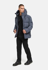 Vadderad grå pufferjacka med huva, framdragkedja och dragsko. Bärs över en svart skjorta och kombineras med svarta byxor och stövlar.