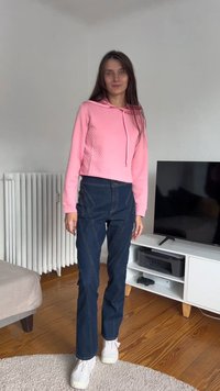 Rosa, kort hoodie med teksturert mønster, kombinert med mørkeblå, høytlivsjeans med kontraststikning. Hvite joggesko fullfører antrekket.