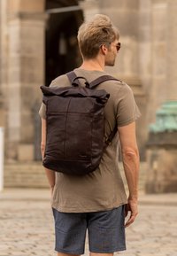 Hnedý kožený batoh s rolovacím vrchom, ktorý má nastaviteľné popruhy, vyznačuje sa prešitým dizajnom a tkaninovou textúrou, uzavretý s hornou rukoväťou.