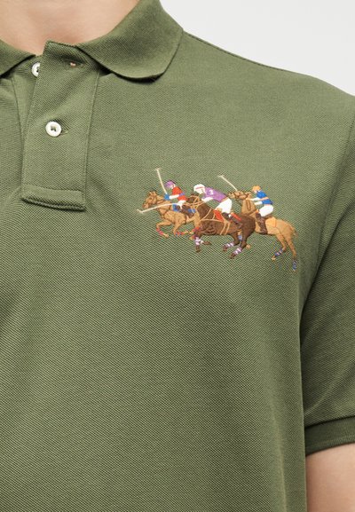 Πράσινο πόλο πουκάμισο με κεντητό σχέδιο τριών παίκτων πόλο που ιππεύουν άλογα, ο καθένας κρατώντας ένα μαλέτι σε μέση κίνηση.