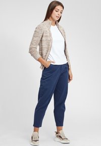 Lys brun strikket zip-jakke over en hvid t-shirt, parret med marineblå tapered bukser og beige sneakers med brune accenter.
