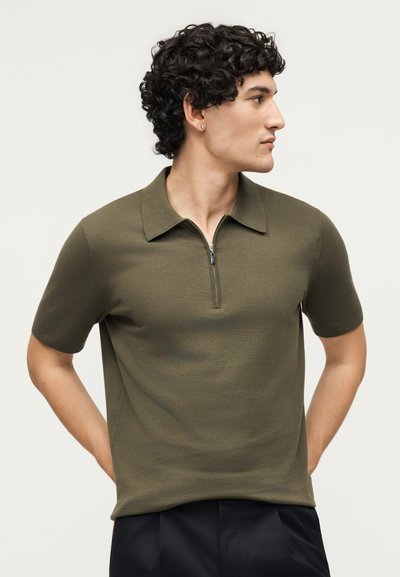 Άνδρας με σγουρά μαλλιά που φοράει χακί κοντομάνικο πόλο με φερμουάρ μπροστά, κοιτάζει πλάγια, τα χέρια πίσω στην πλάτη, με φόντο ένα απλό ανοιχτόχρωμο φόντο.